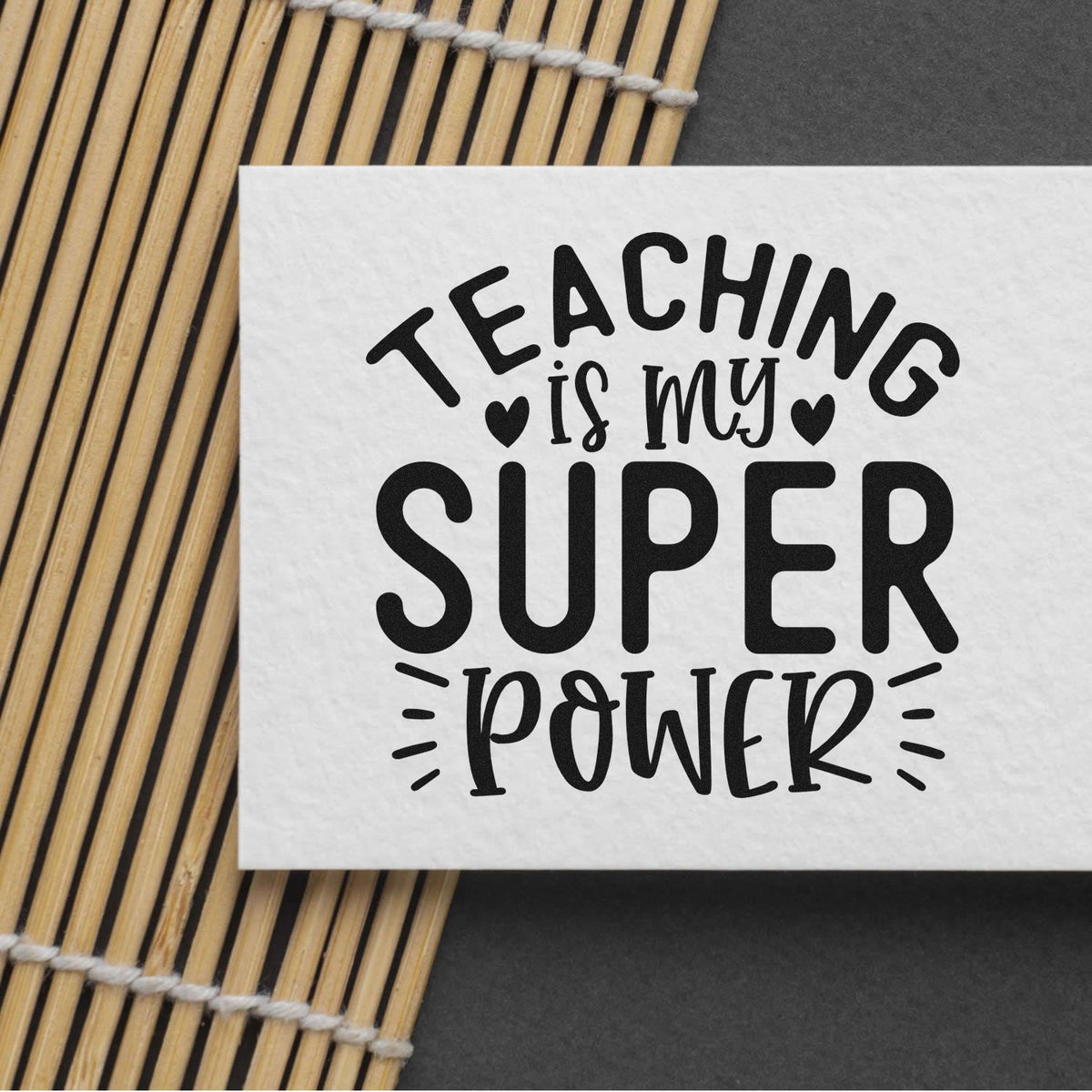 Black teaching stamp/seal on white paper reads Teaching is my Super Power, with bamboo mat on dark background.