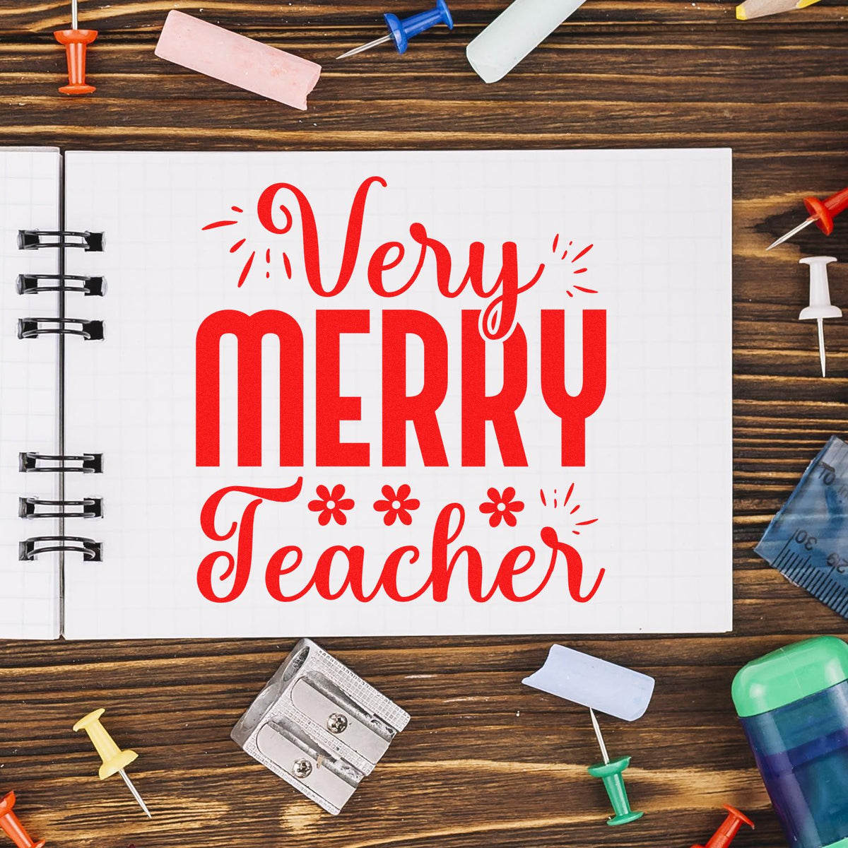 Red Very Merry Teacher stamp and seal design on a white notebook, surrounded by colorful school supplies on wood.