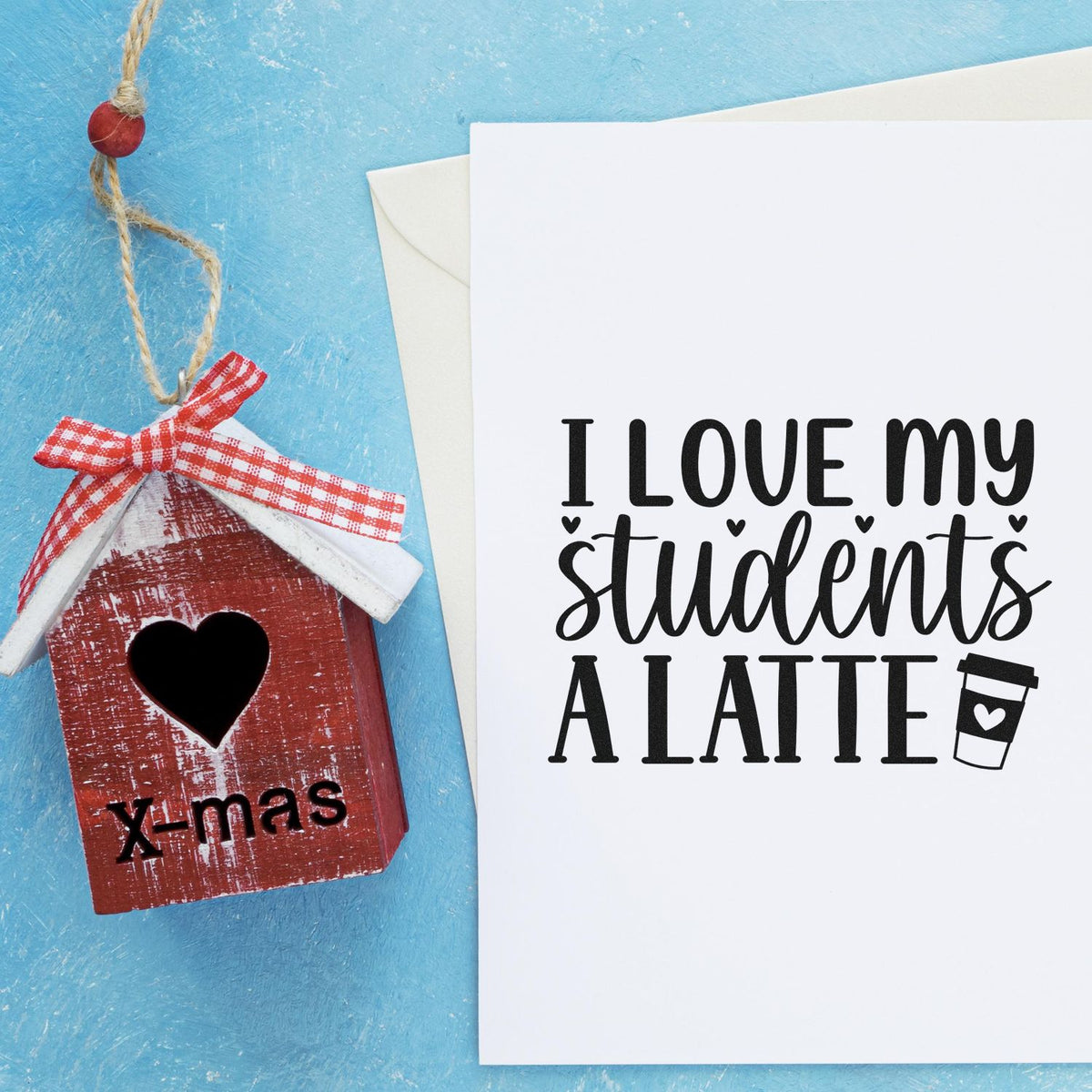 Red birdhouse stamp seal with heart cutout and plaid ribbon beside white card reading I love my students a latte on blue background.