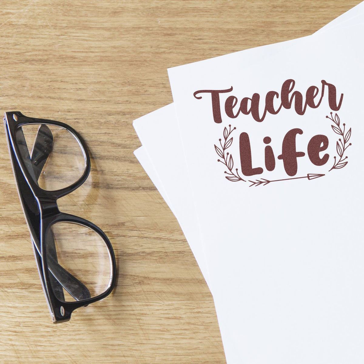 Black eyeglasses beside white paper with brown Teacher Life stamp and seal design on wood desk