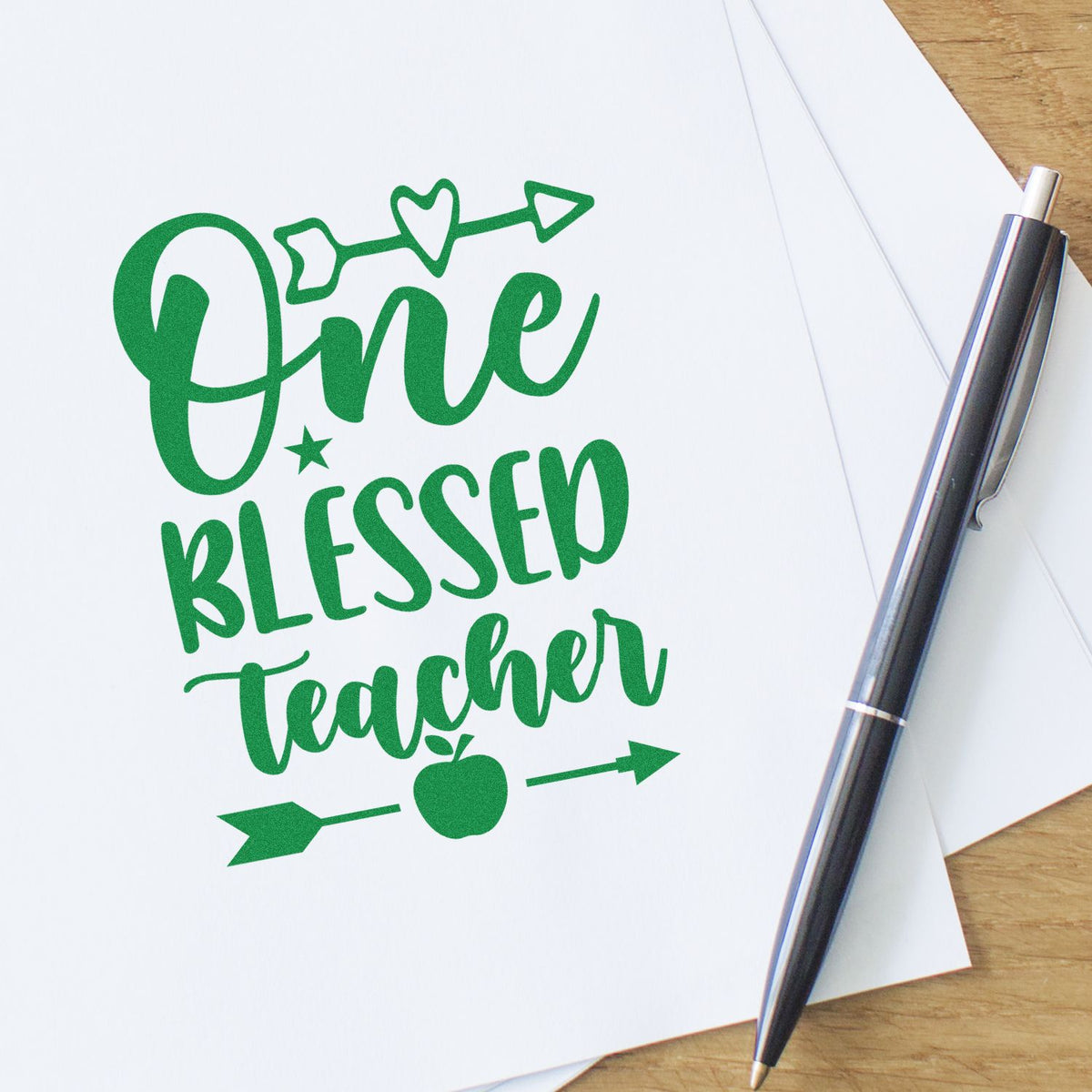 Green One Blessed Teacher stamp seal design with heart, star, apple and arrows beside a silver pen on white paper.