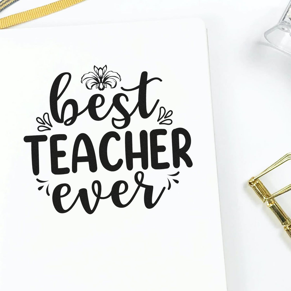 Black Best Teacher Ever stamp seal on white paper, with floral accents and gold clips