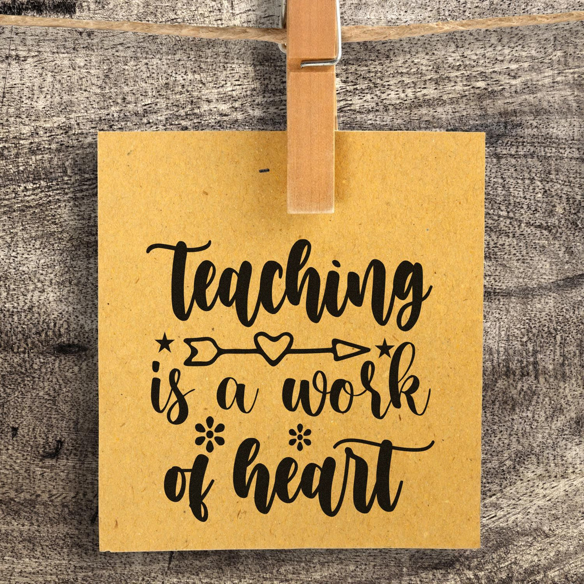 Tan hanging stamp seal card with black calligraphy reading Teaching is a work of heart, clipped to rustic wood.