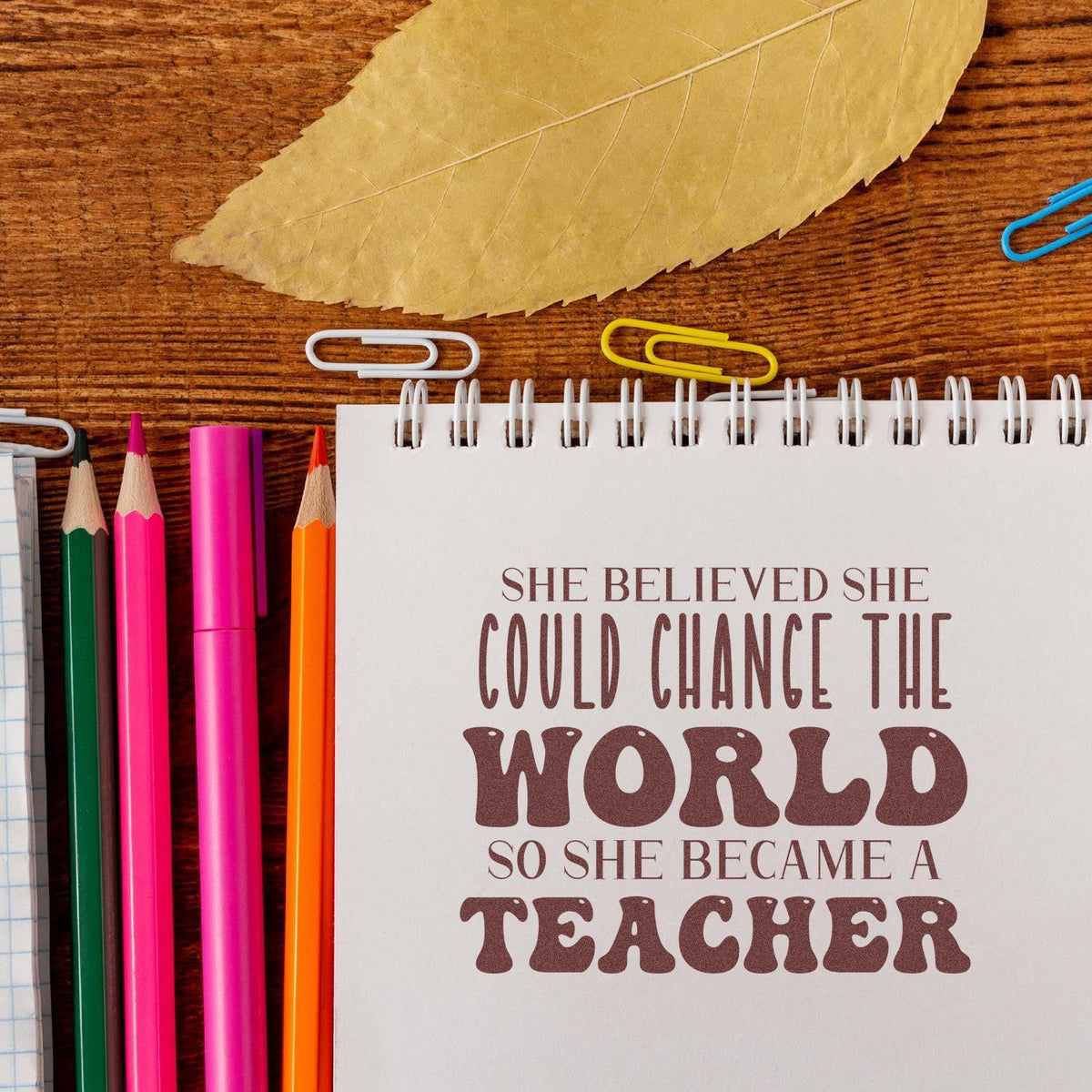 Custom stamp and seal design on a white spiral notebook with brown quote, colorful pencils, paper clips, and a tan leaf on wood.