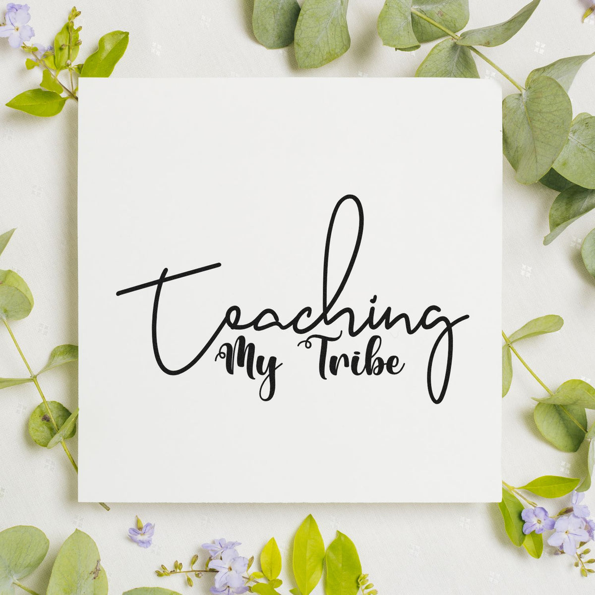 Custom stamp and seal with black Teaching My Tribe script on a white card, surrounded by green leaves and lavender flowers.