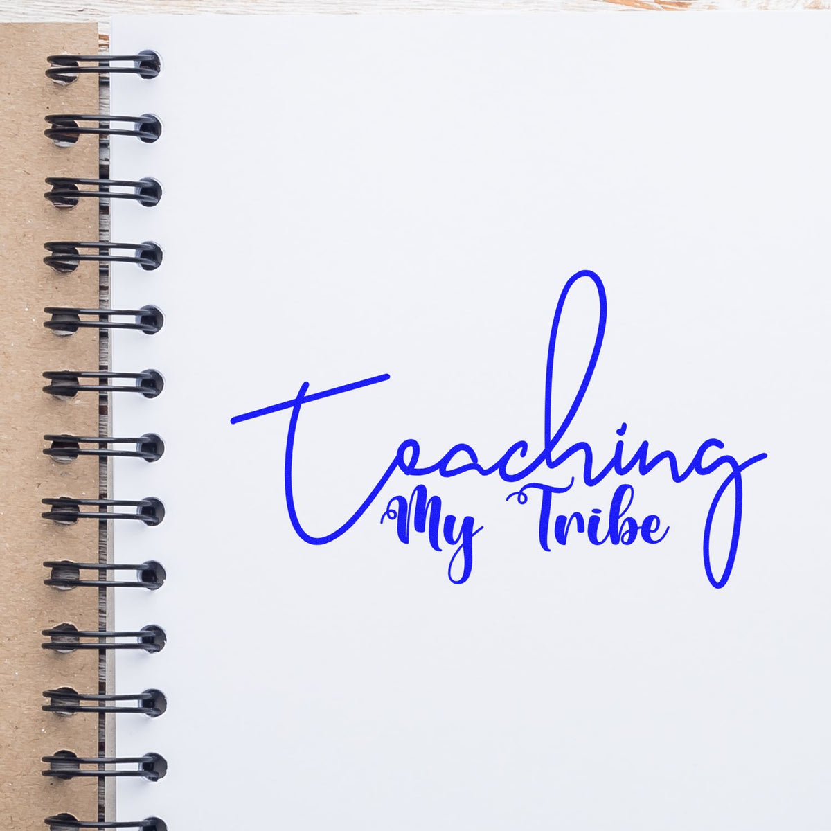Blue Teaching My Tribe stamp and seal design on a white spiral notebook with kraft cover and black metal binding.