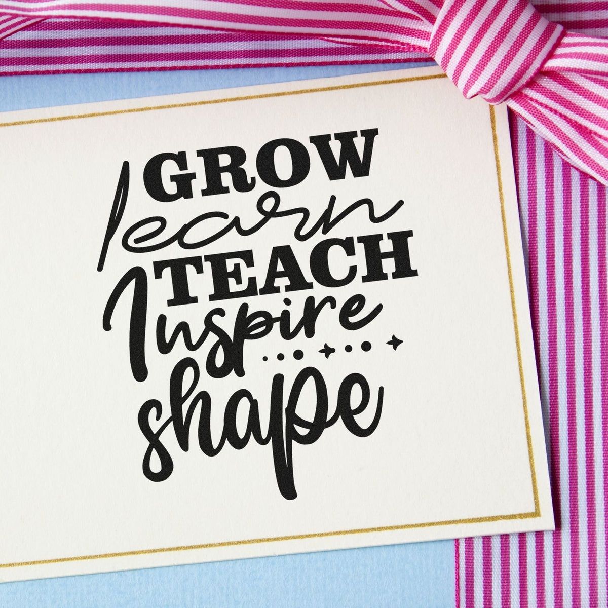 Black custom stamp reading Grow learn teach inspire shape on cream card with gold border and pink striped ribbon.