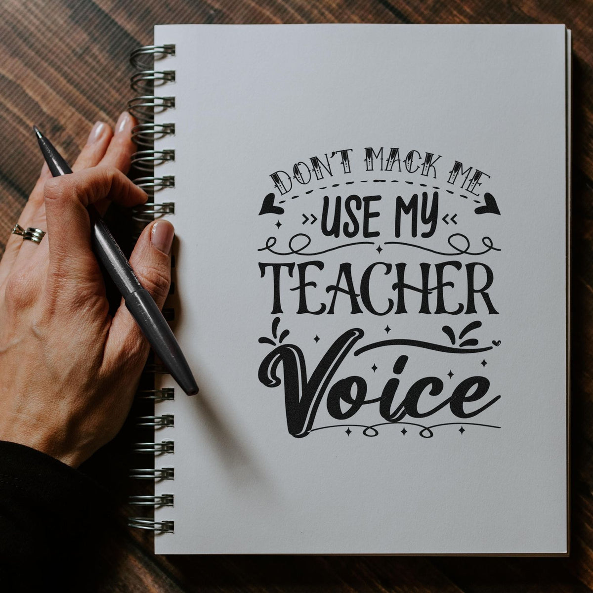 Black teacher-voice stamp/seal design on a white spiral notebook, with a hand holding a pen.