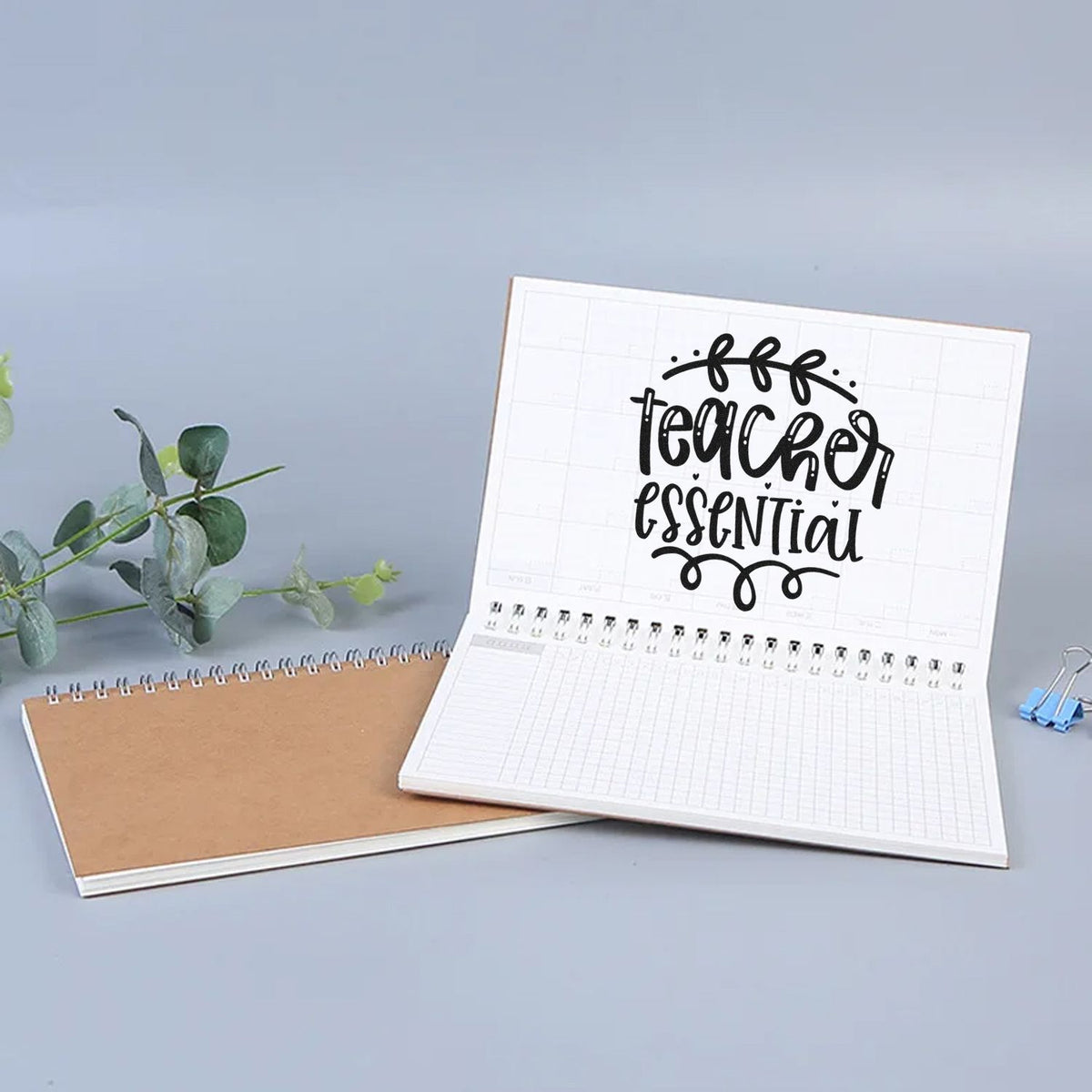 Black Teacher Essential stamp on open white planner notebook, beside a kraft spiral notebook, green leaves, and a blue binder clip.