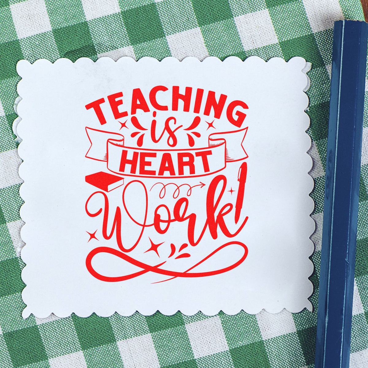 Red decorative teaching stamp reading Teaching is Heart Work on a white scalloped seal over green gingham with blue pen.