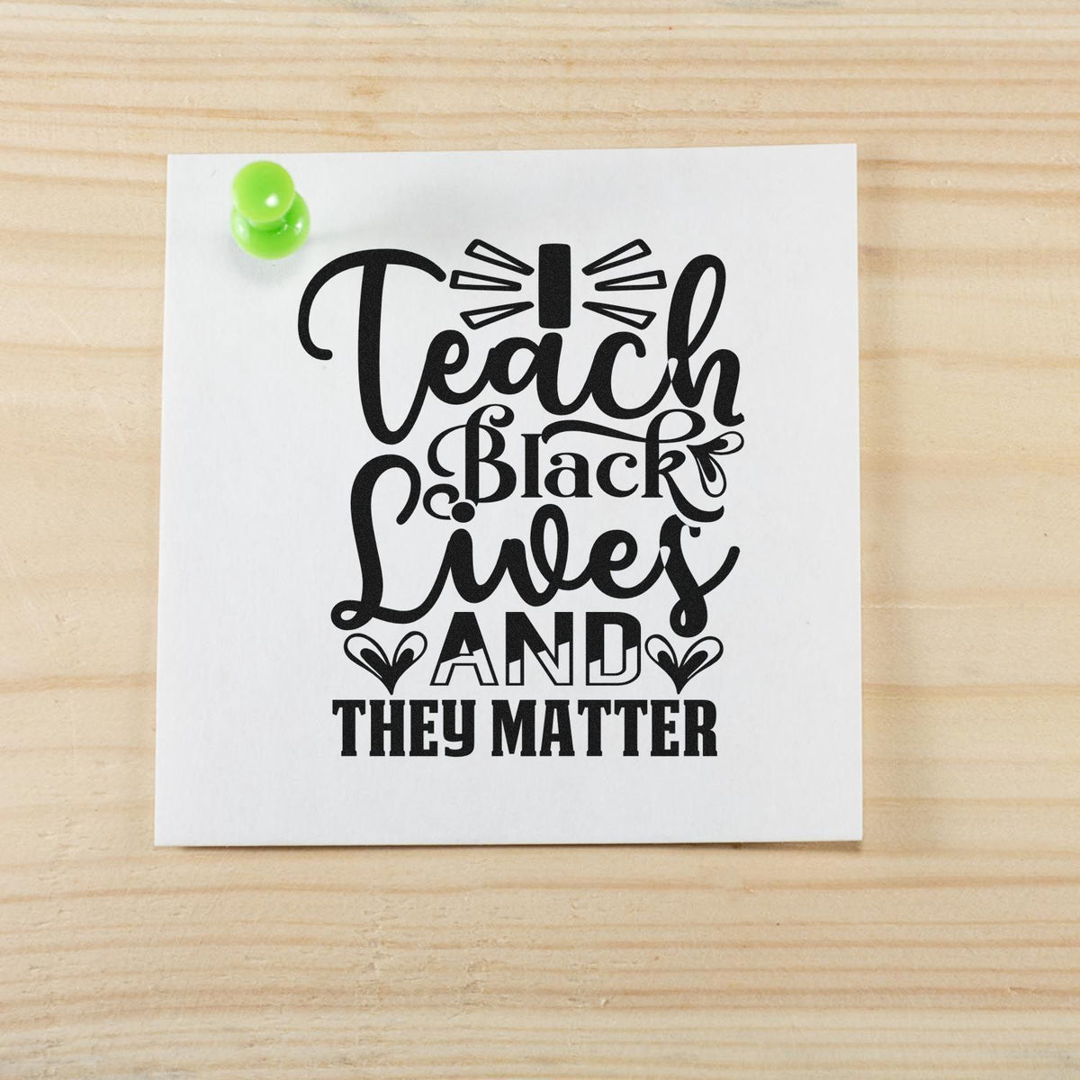 Black typographic stamp design reading Teach Black Lives and They Matter on a white card, pinned with a green pushpin.
