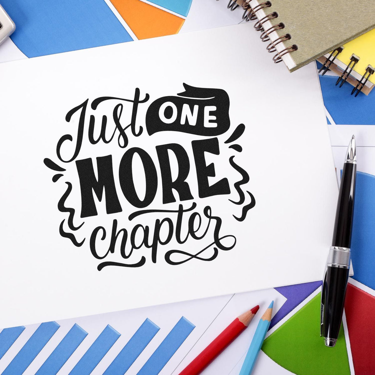 Black custom stamp and seal reading Just One More Chapter on white paper, with colorful envelopes, pens, and notebooks.
