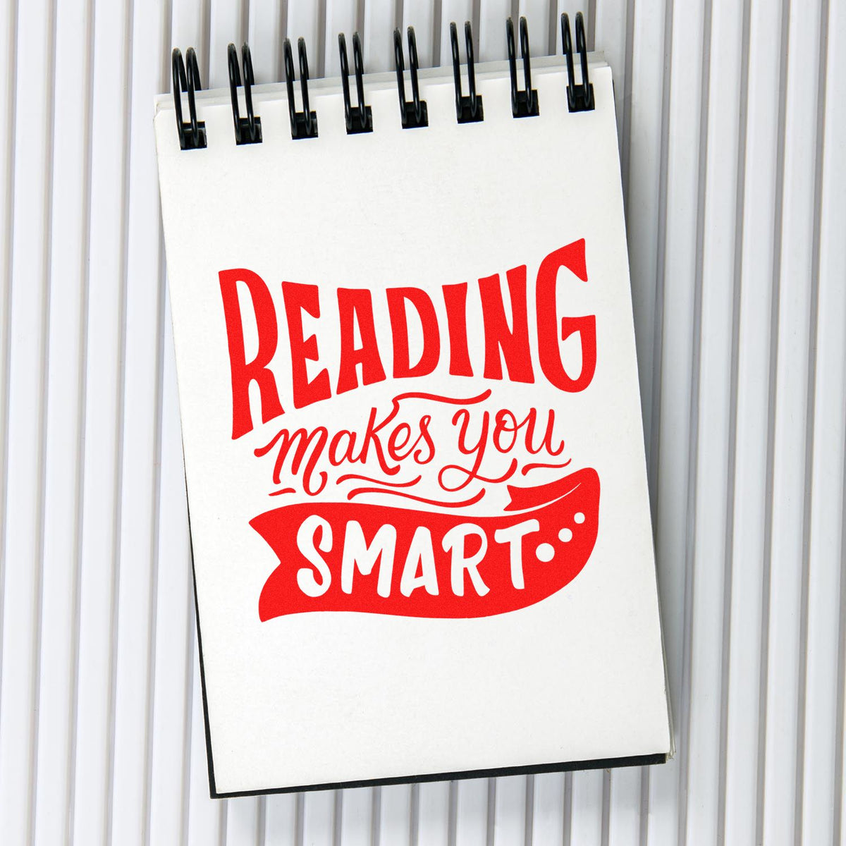 White spiral notepad with bold red stamp-and-seal style text reading Reading makes you smart on a gray striped background.