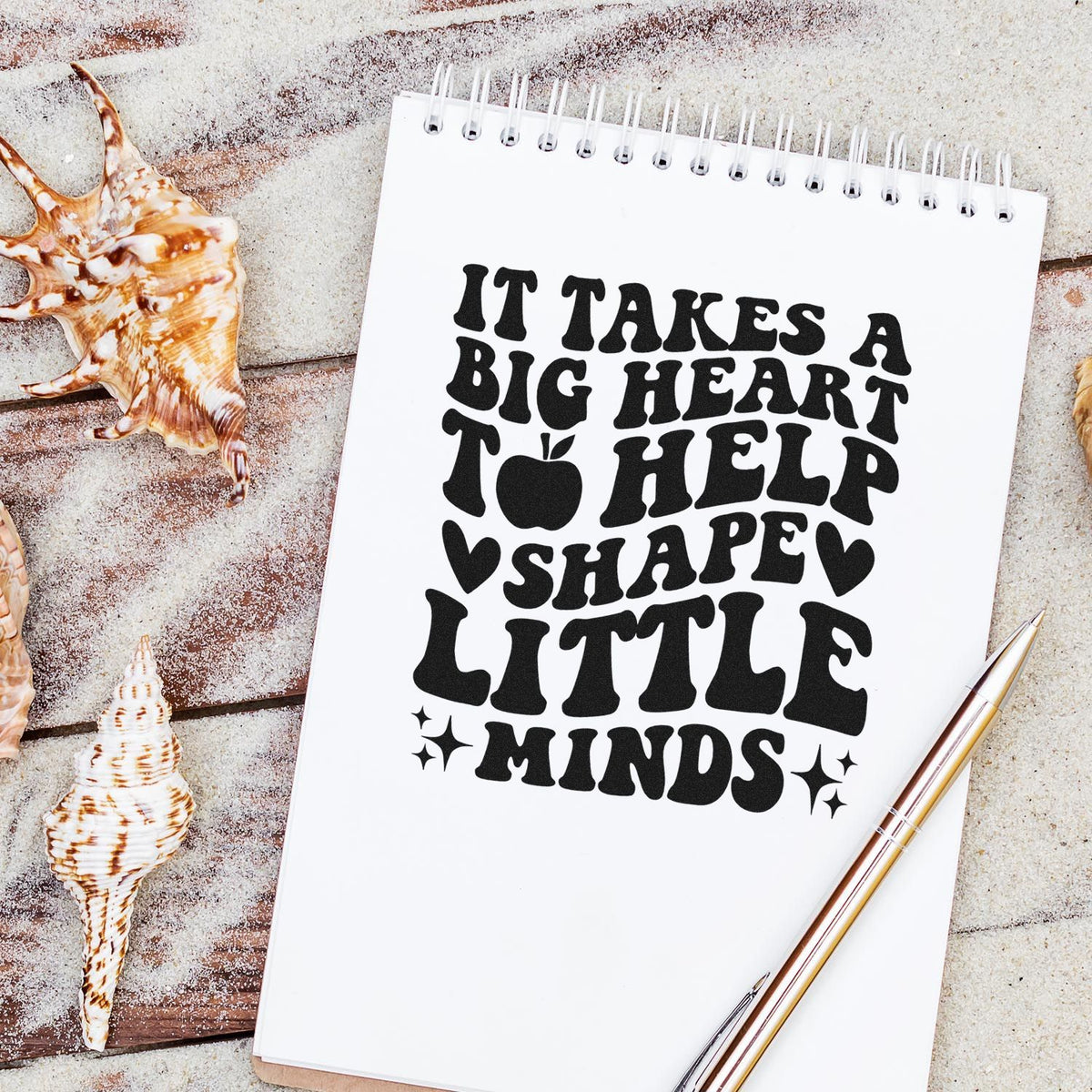 Black stamp-style quote on white spiral notebook: It takes a big heart to help shape little minds, with seashells and pen.