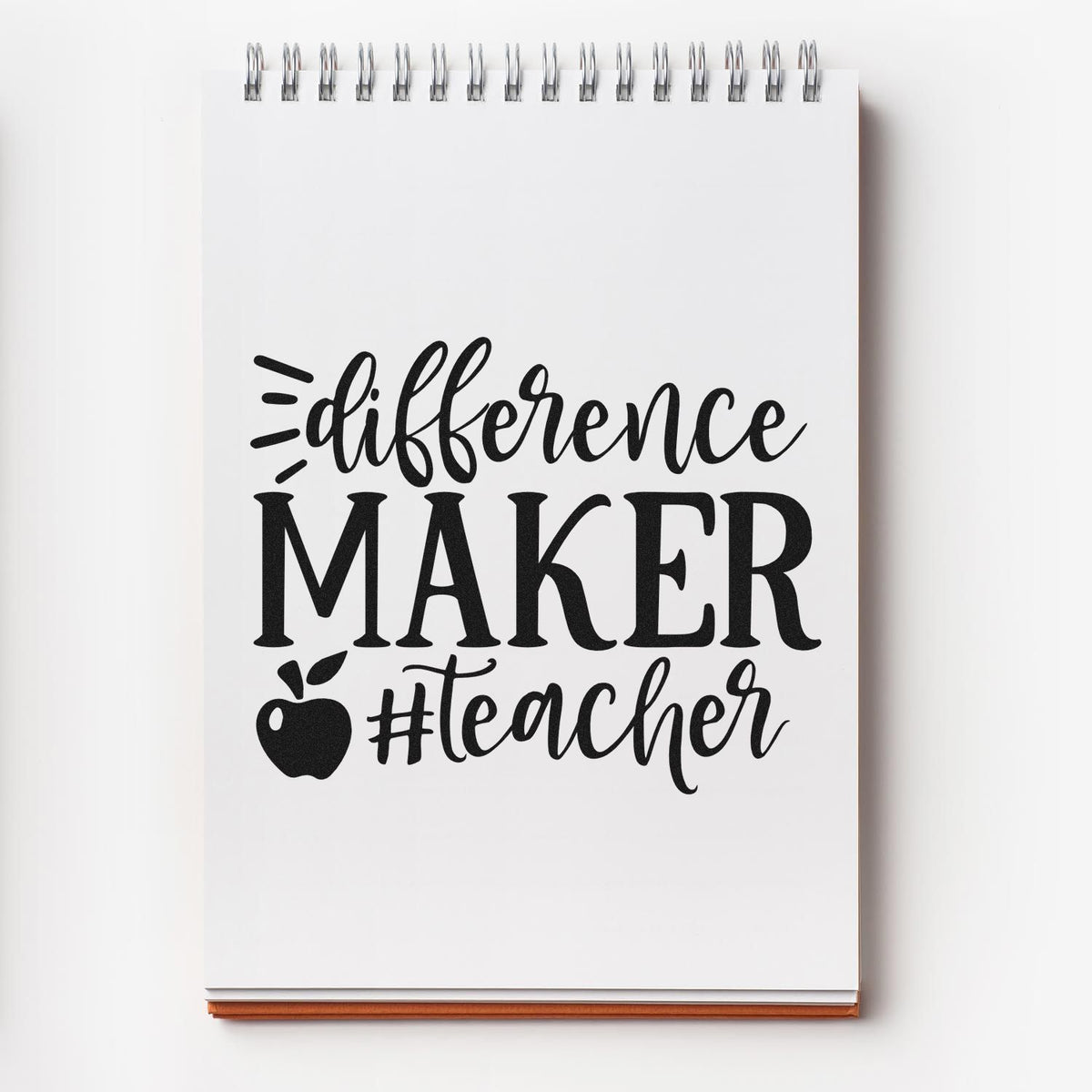Black teacher stamp and seal design on a white spiral notebook, with apple icon and bold difference MAKER #teacher text.