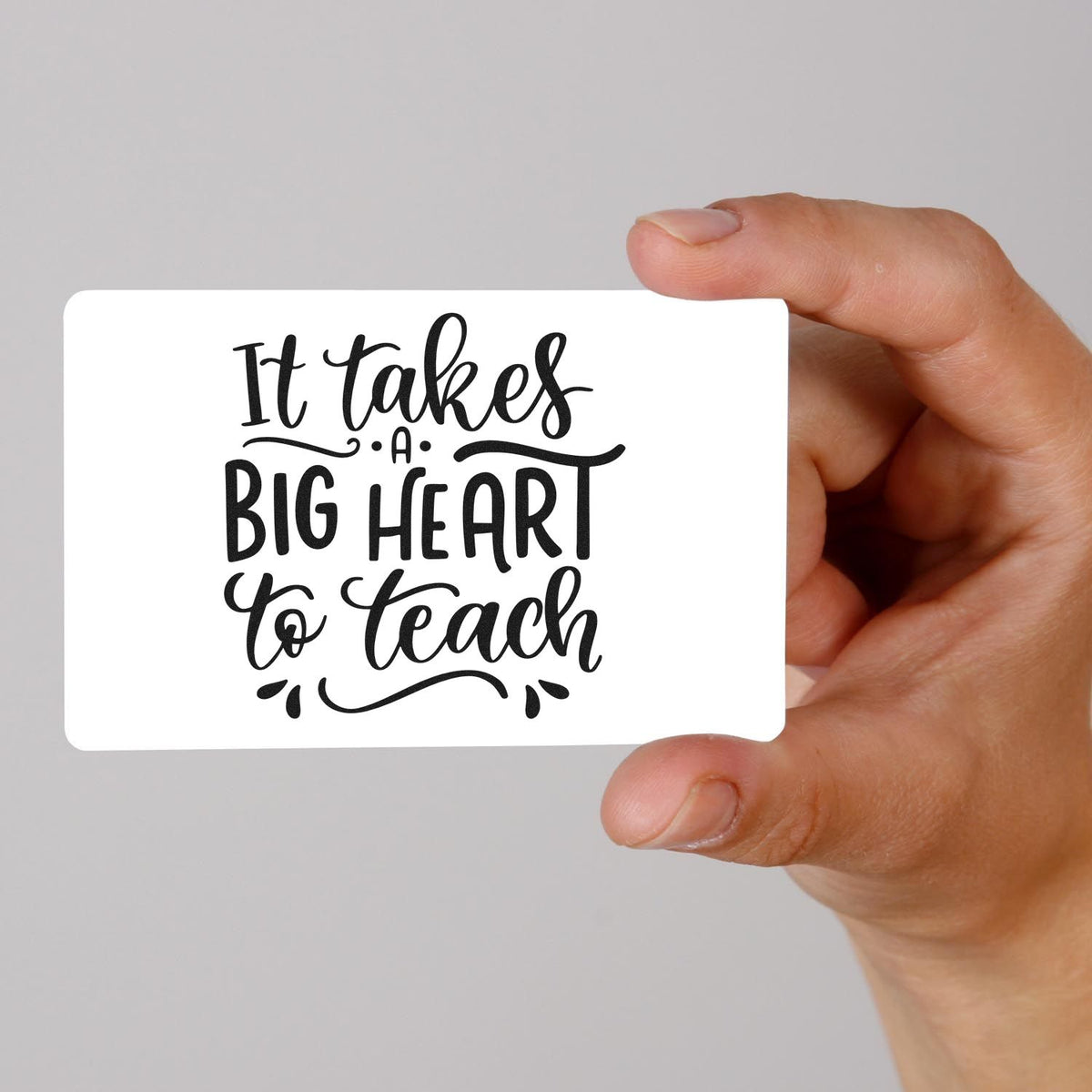 Hand holding a white stamp and seal sample card with black It takes a big heart to teach lettering on gray.
