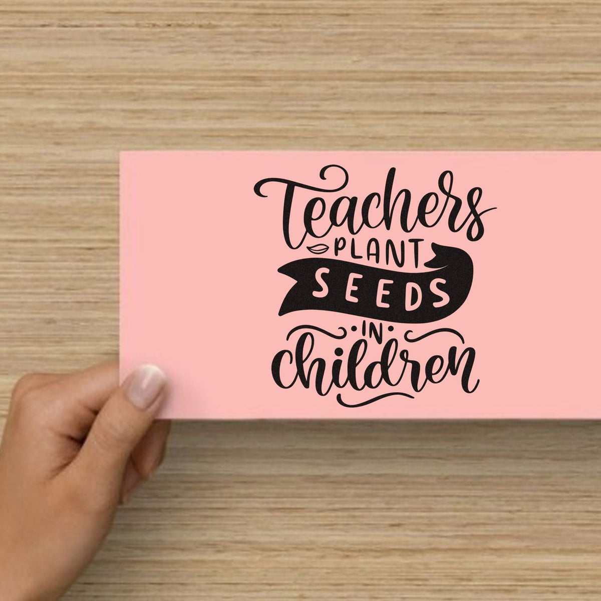 Pink stamp and seal card with black Teachers plant seeds in children lettering on a wood background.