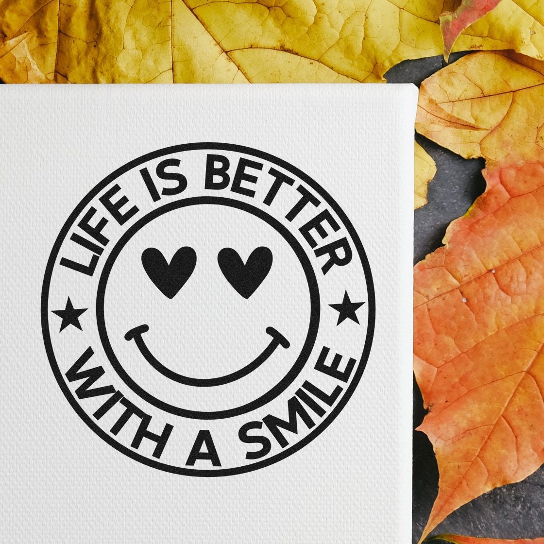 Black round rubber stamp seal with heart-eyes smiley and Life is Better With a Smile on white paper, autumn leaves background.