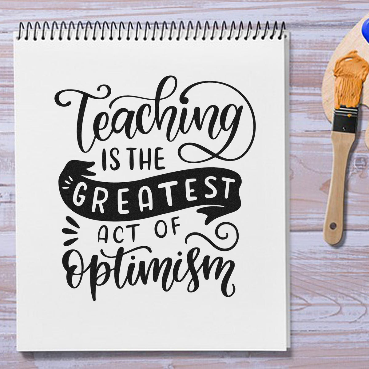 Black teaching quote on white spiral notebook beside a wooden stamp brush, on a light wood background.