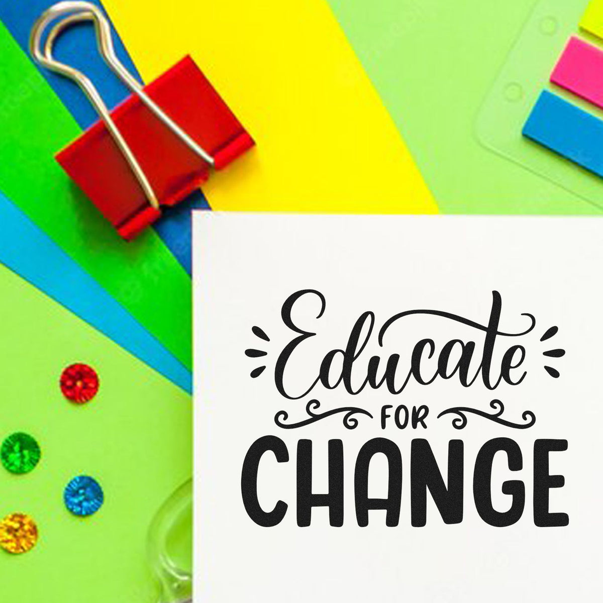 White stamp card reading Educate for Change on colorful paper background with red binder clip and glitter accents