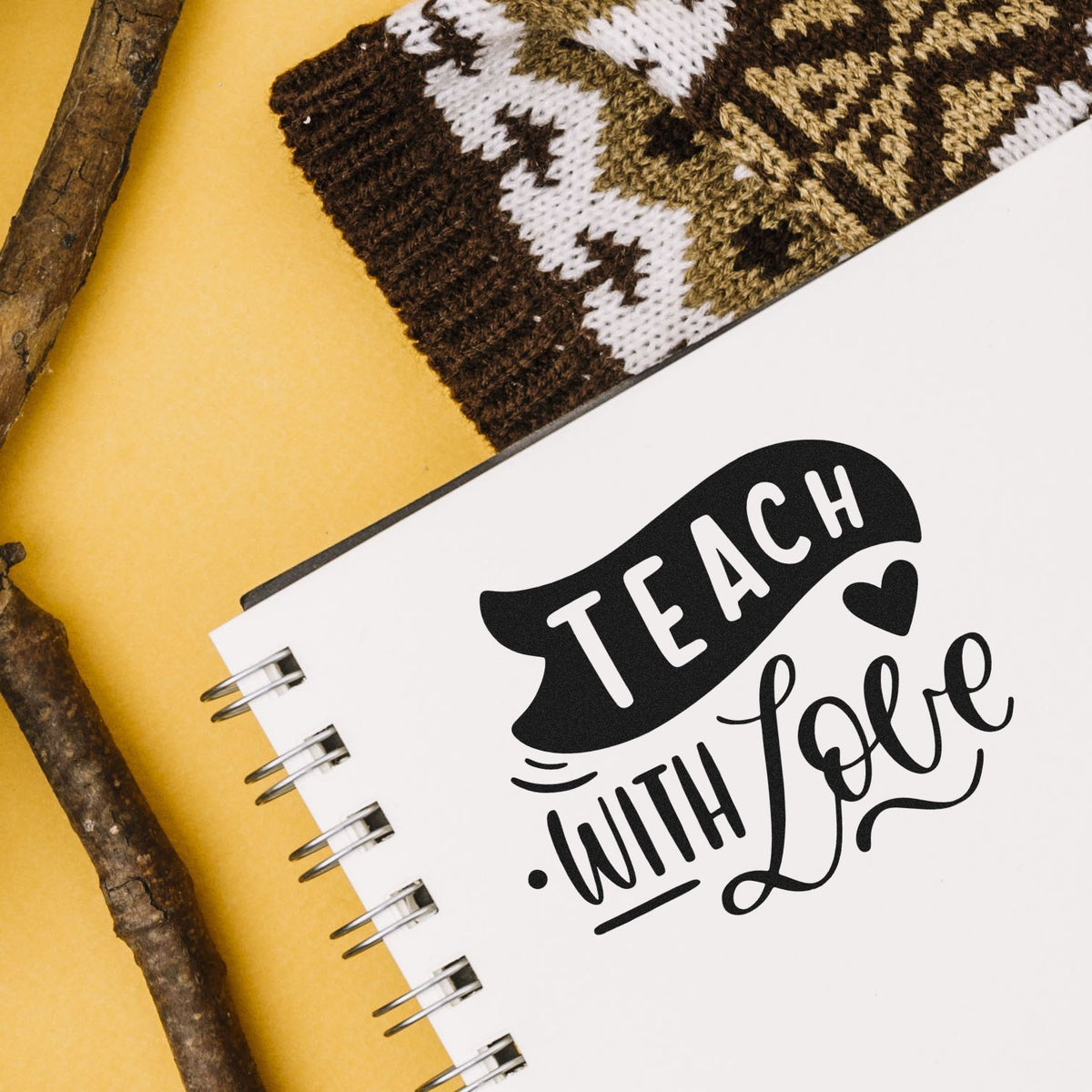 Black Teach with Love stamp and seal on white spiral notebook, on mustard background with knit fabric and twigs.