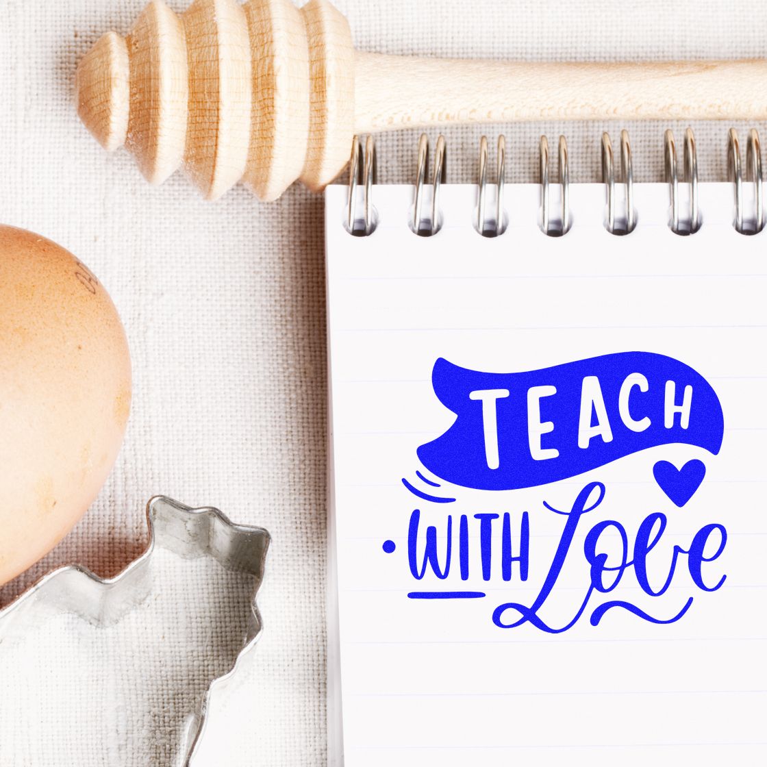 Blue Teach with Love stamp on white spiral notebook, beside wooden stamp handle, egg, and silver cookie cutter.