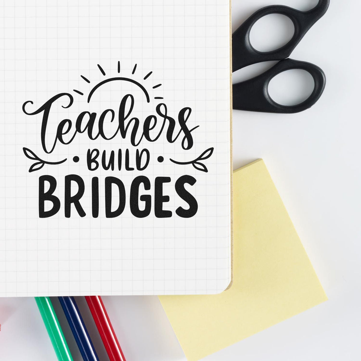 Black teacher stamp seal reading Teachers Build Bridges on grid paper, with scissors, yellow note, and colorful pens.