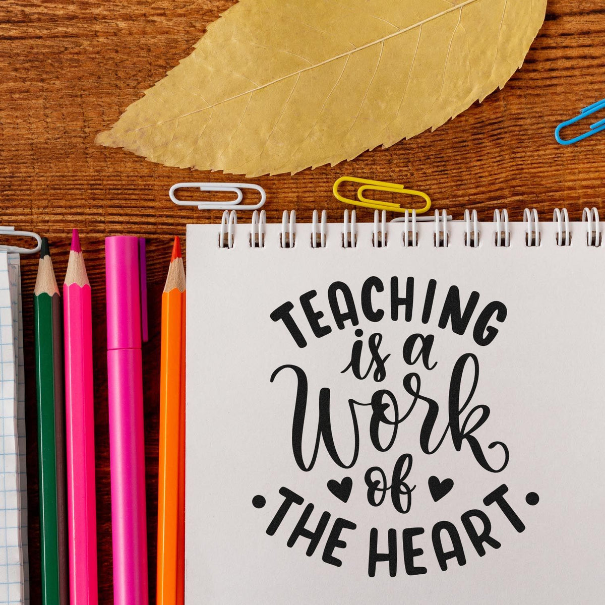 Decorative teaching quote stamp/seal on white spiral notebook, with pink, orange, and green pencils, paper clips, and tan leaf.