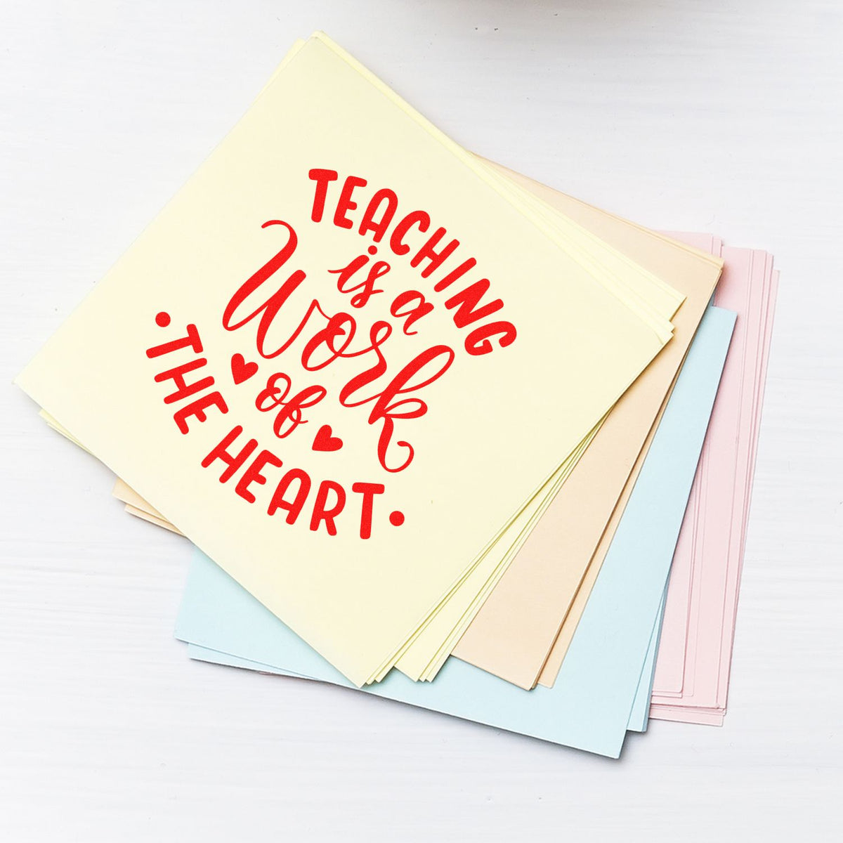 Red rubber stamp reading Teaching is a Work of the Heart on cream cardstock, with pastel blue, pink, beige sheets.