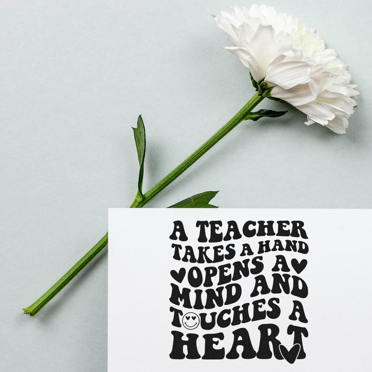 White flower with green stem beside a white card featuring a black teacher stamp and seal design with heart accents.