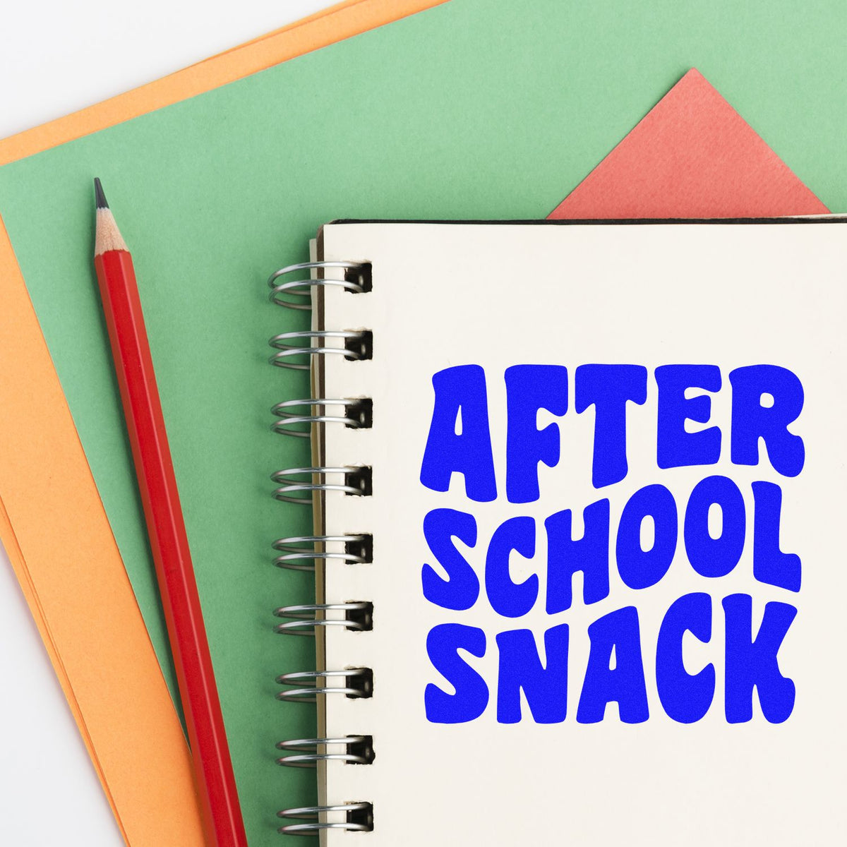 Blue AFTER SCHOOL SNACK custom stamp on cream spiral notebook, with red pencil, green background, and orange paper.