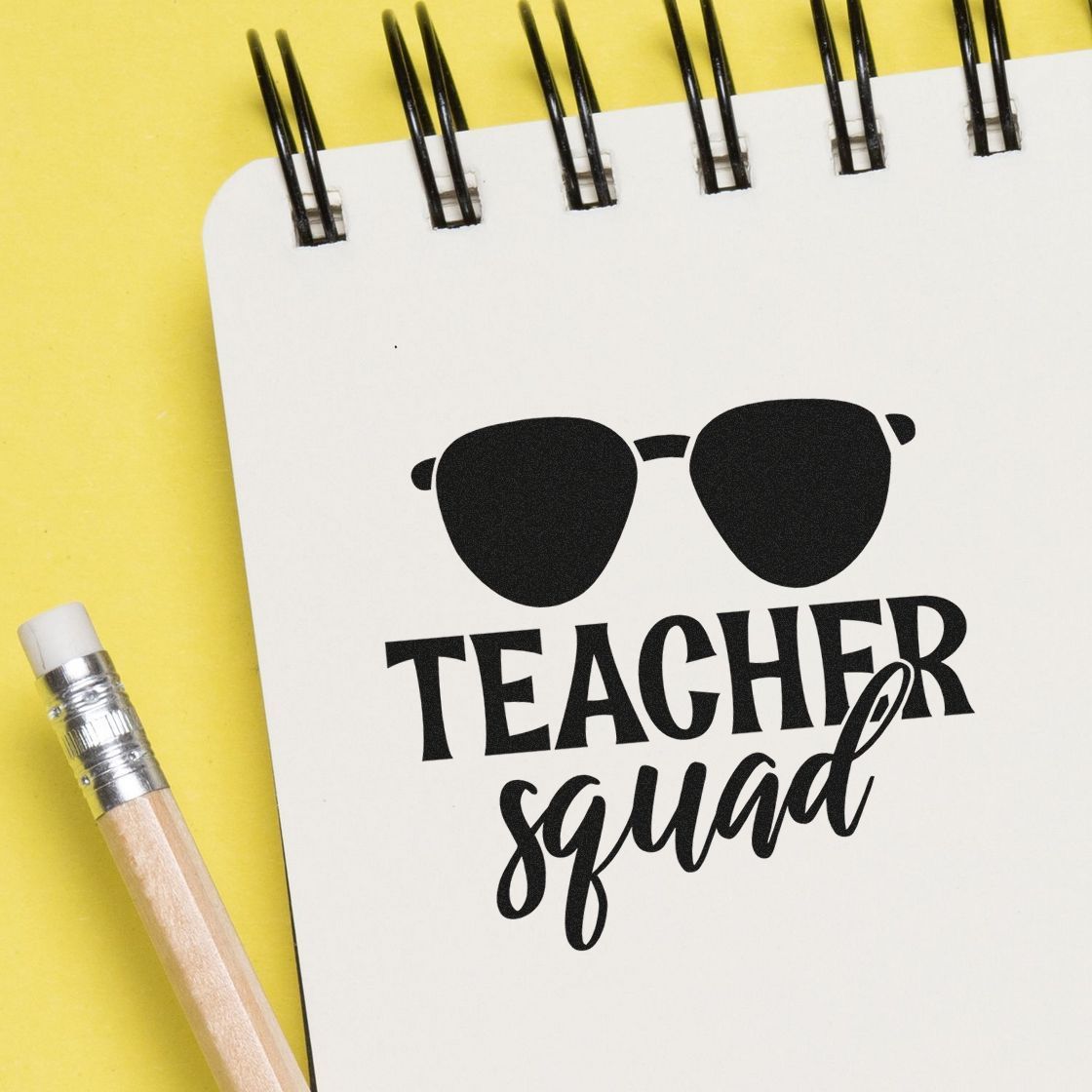 Black Teacher Squad stamp seal with sunglasses on white notebook, beside a pencil on a yellow background.