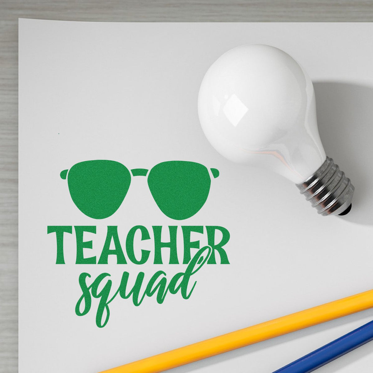 Green Teacher Squad stamp and seal design in green, with sunglasses icon on white paper beside a light bulb and colored pencils.