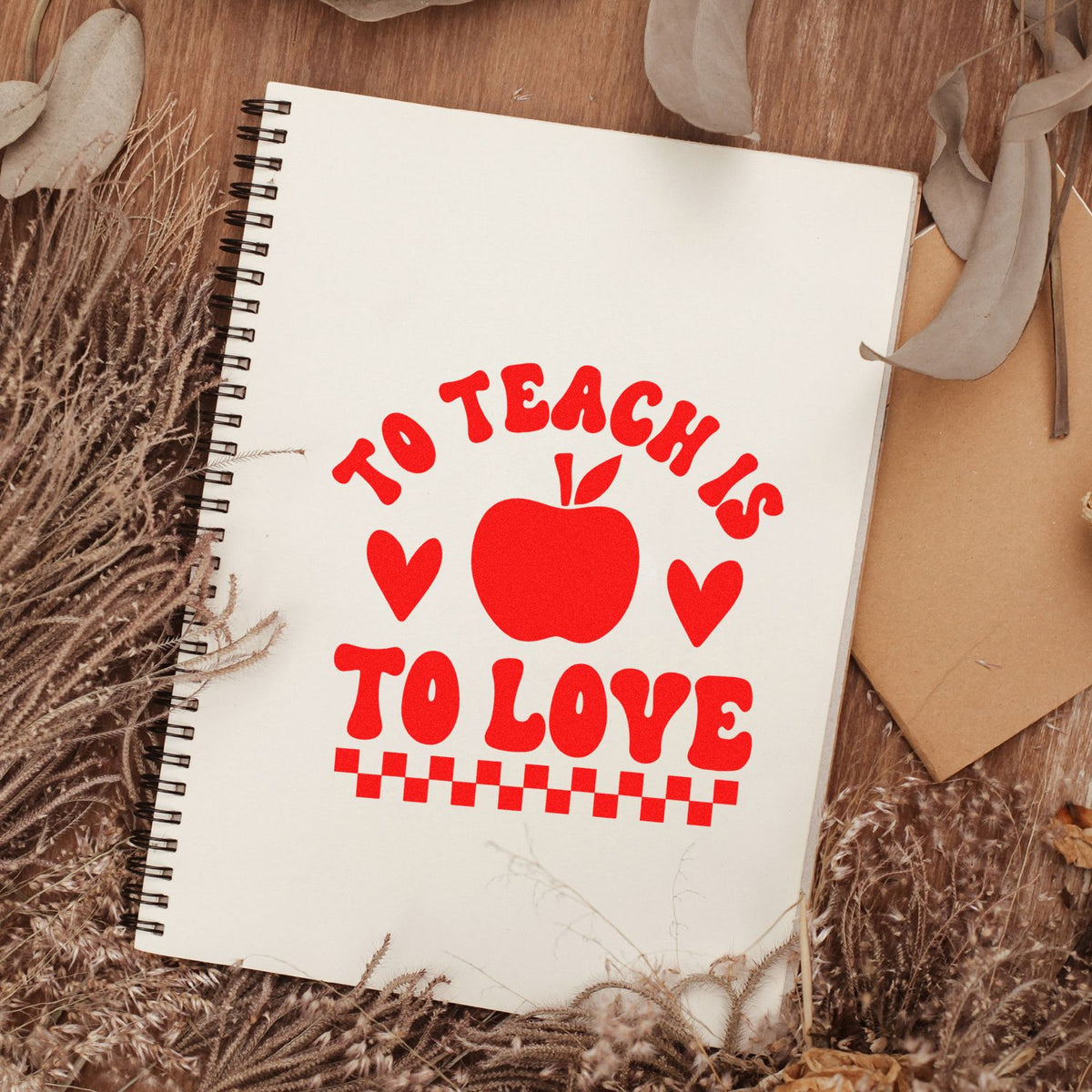 Red teacher stamp seal with apple and hearts on a white spiral notebook, surrounded by dried florals.
