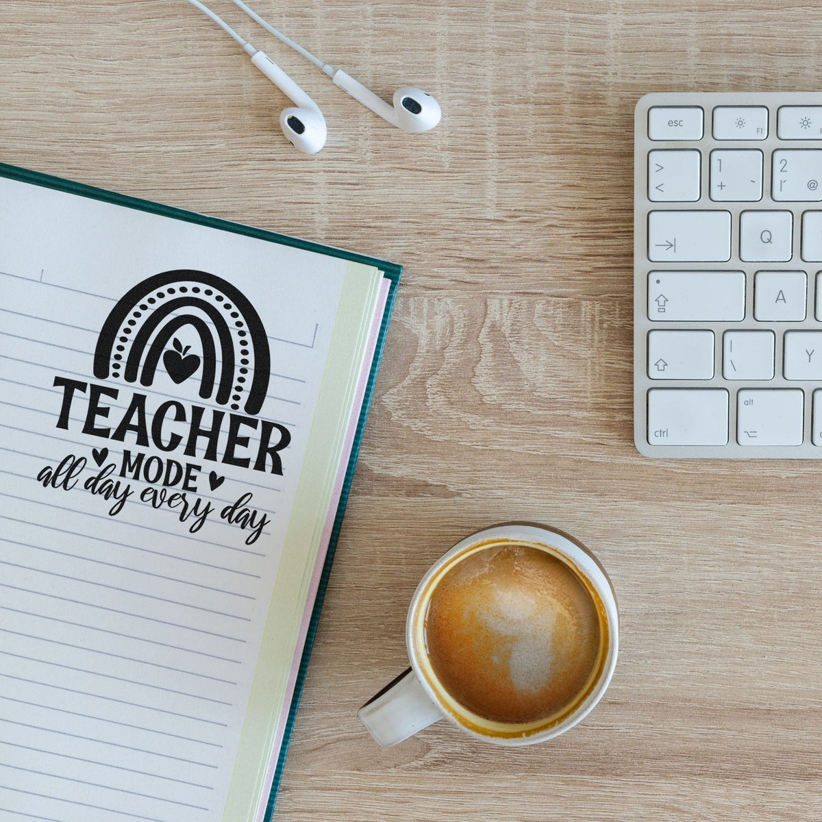 Black Teacher Mode stamp and seal on lined notebook, beside keyboard, earbuds and coffee on a light wood desk.