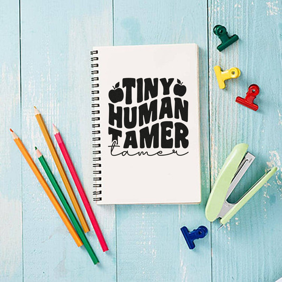 Tiny Human Tamer stamp design on a white spiral notebook, with colorful pencils, clips, and a mint stapler on blue wood.