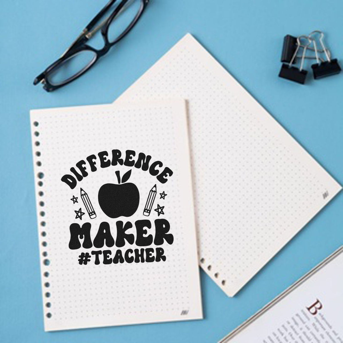 Black Difference Maker #Teacher stamp seal with apple and pencils on white dotted pages over a blue background.