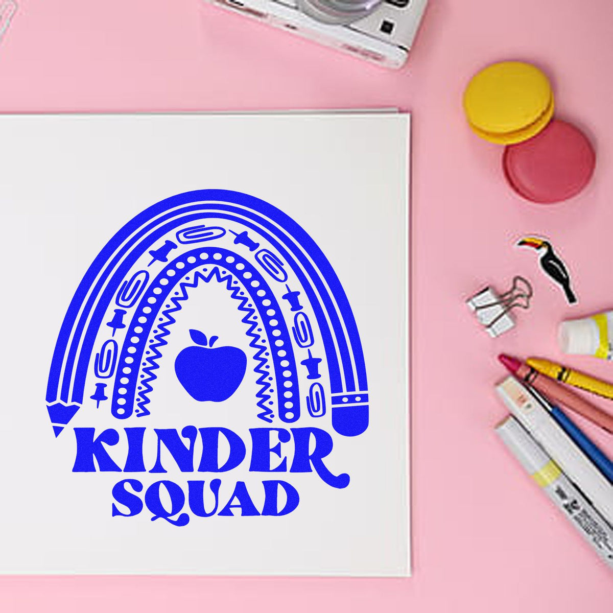 Blue Kinder Squad rainbow apple stamp design on a white card, with pink background and colorful crafting tools and seal pads nearby