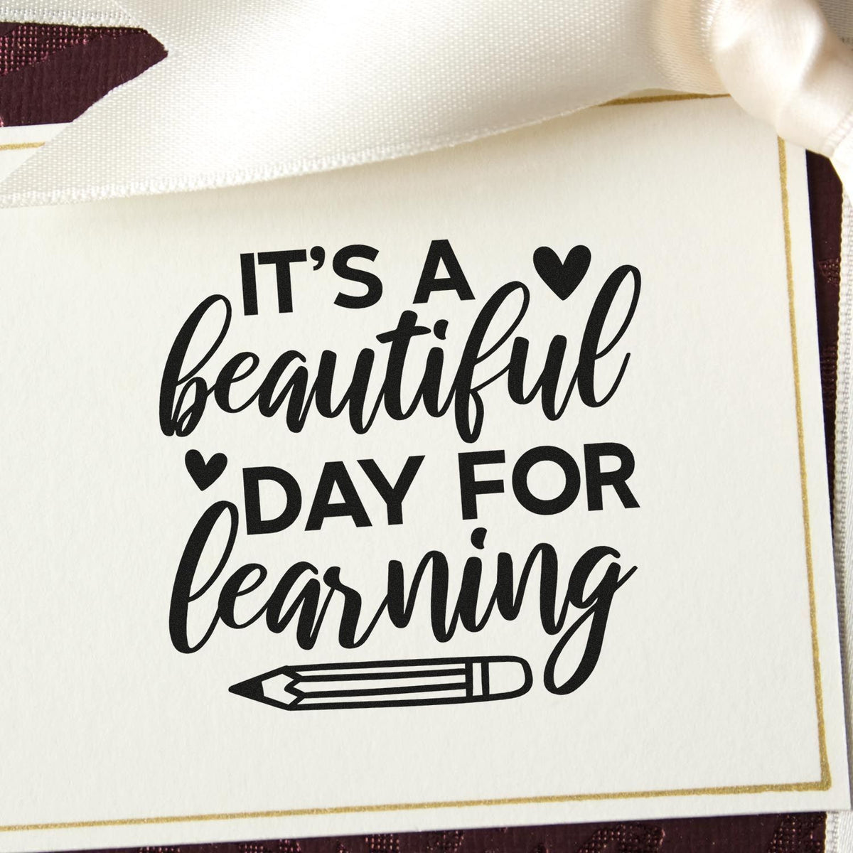 White stamp seal with black Its a beautiful day for learning text, hearts, pencil icon, gold border, and satin ribbon.
