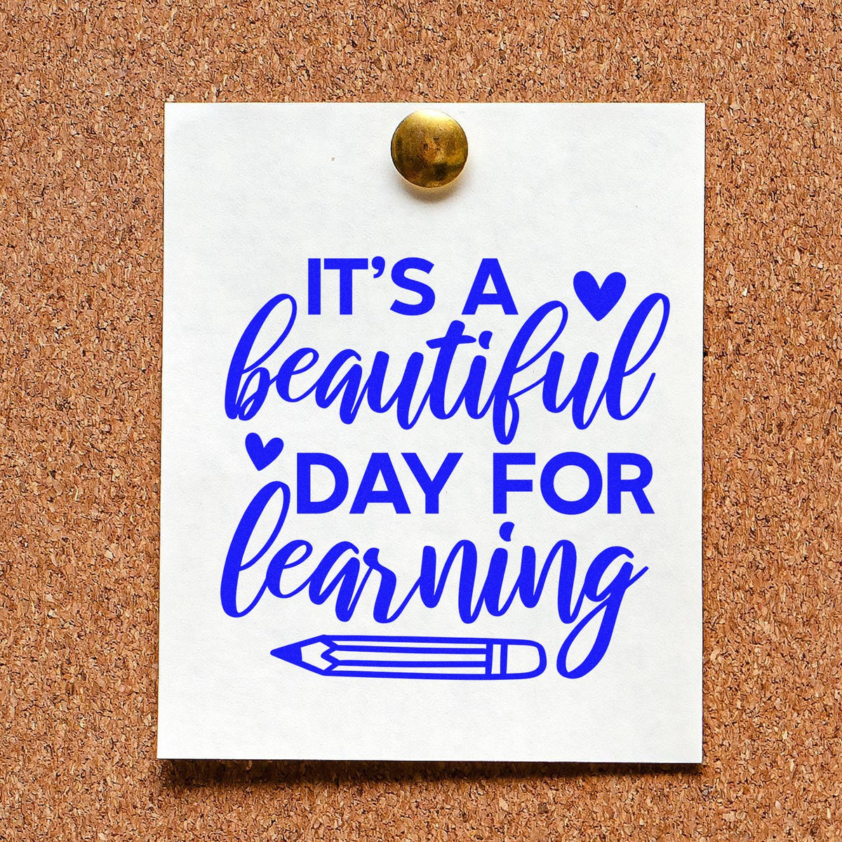 Blue rubber stamp seal on white paper pinned to corkboard: Its a beautiful day for learning with hearts and pencil icon.