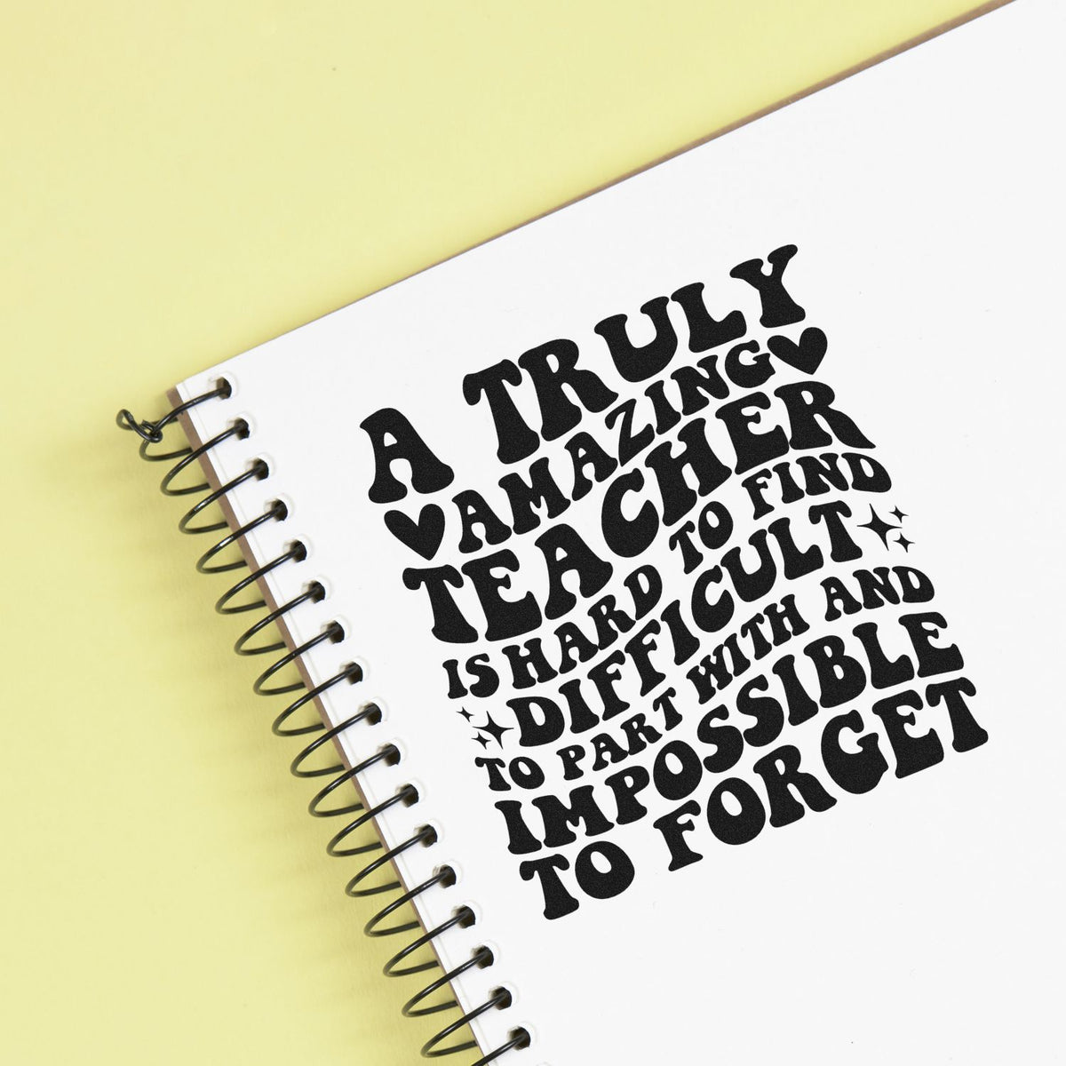 Black teacher appreciation stamp seal on a white spiral notebook, with bold quote, heart, sparkles, on pale yellow background.