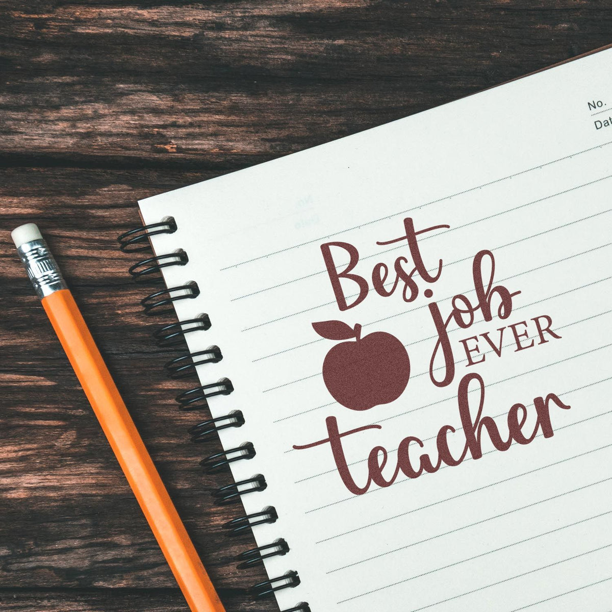 Maroon Best Job Ever teacher stamp with apple icon on a white lined notebook beside an orange pencil on dark wood.