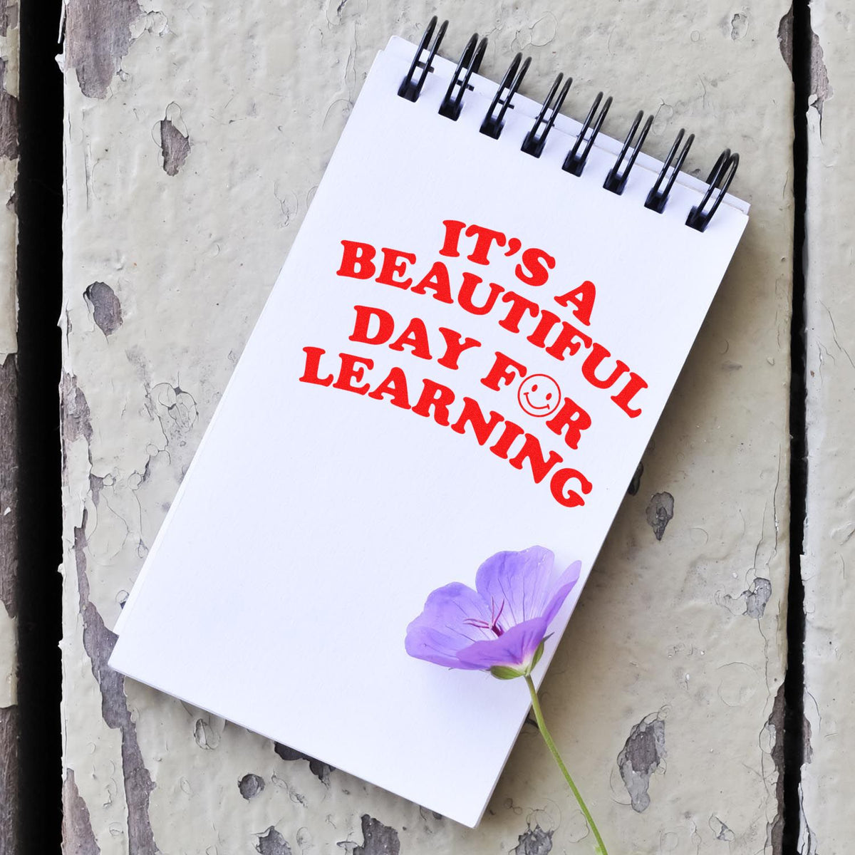 White spiral notebook with bold red stamp-style text, purple flower accent, and black rings on a weathered gray surface.