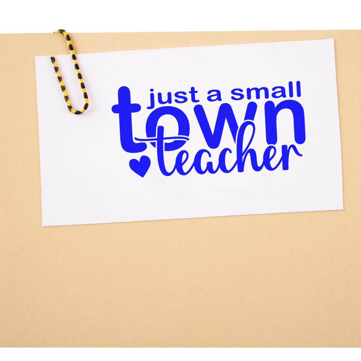 Blue custom stamp design reading just a small town teacher on white card, clipped with yellow-and-black paperclip.