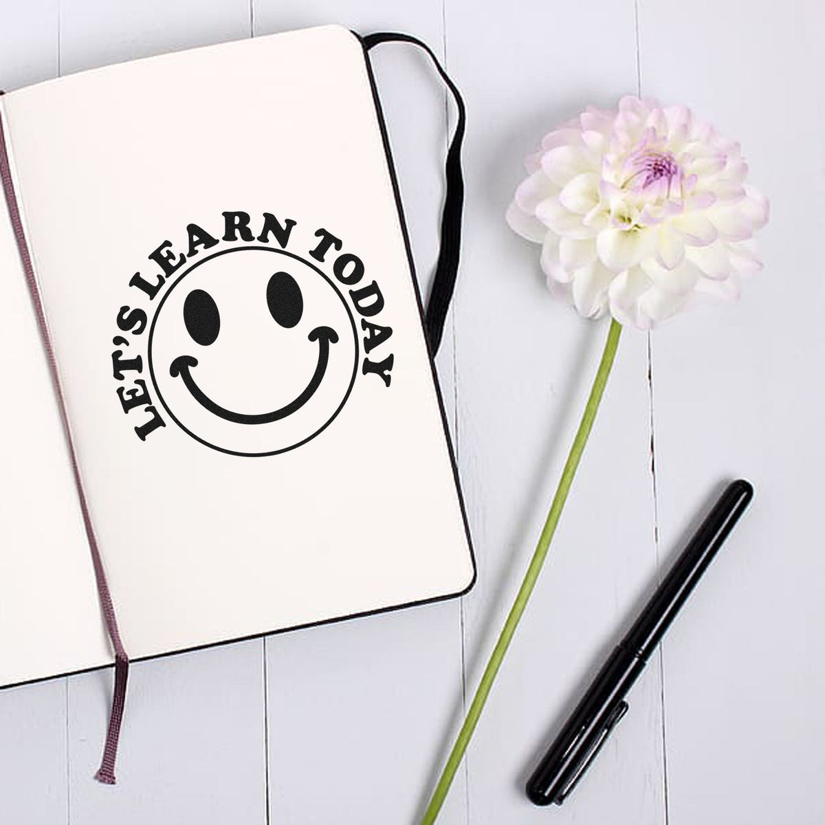 Cream notebook with black Lets Learn Today smiley stamp seal, black pen, and white flower on a white desk.