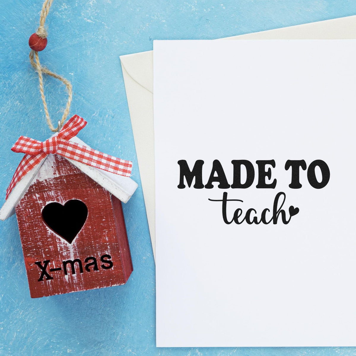 Red birdhouse stamp seal with heart cutout and plaid bow beside white card reading MADE TO teach on blue background.