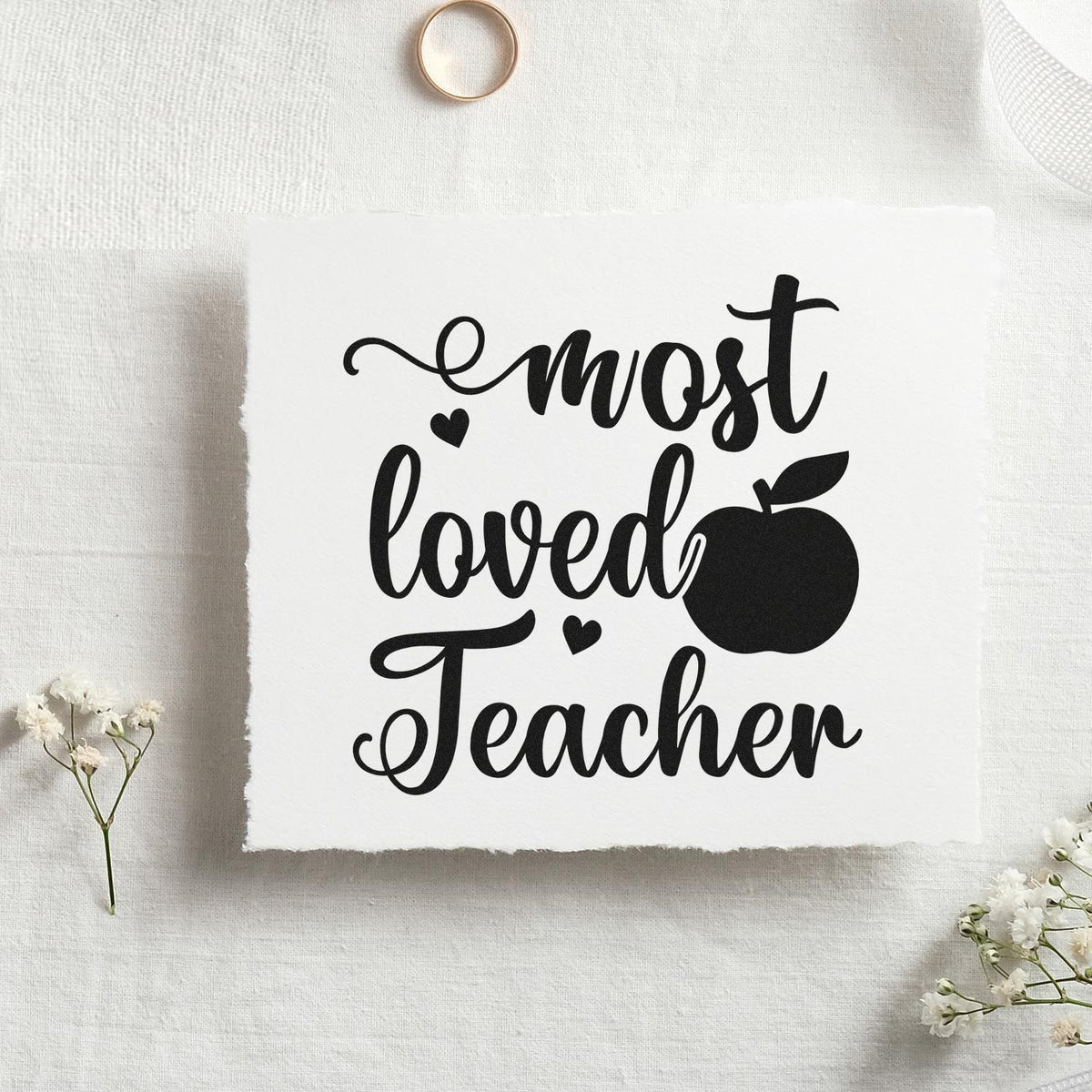 Black most loved Teacher stamp and seal design with apple icon on white textured paper, accented by white flowers.