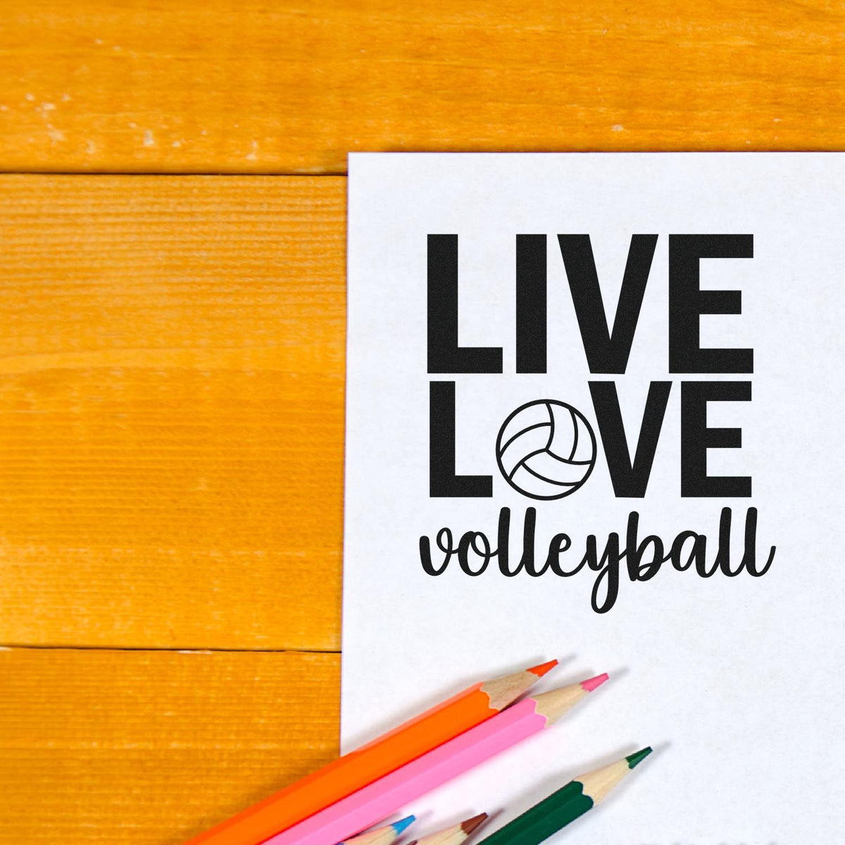 Black Live Love volleyball stamp and seal on white paper, with orange wood background and colorful pencils.