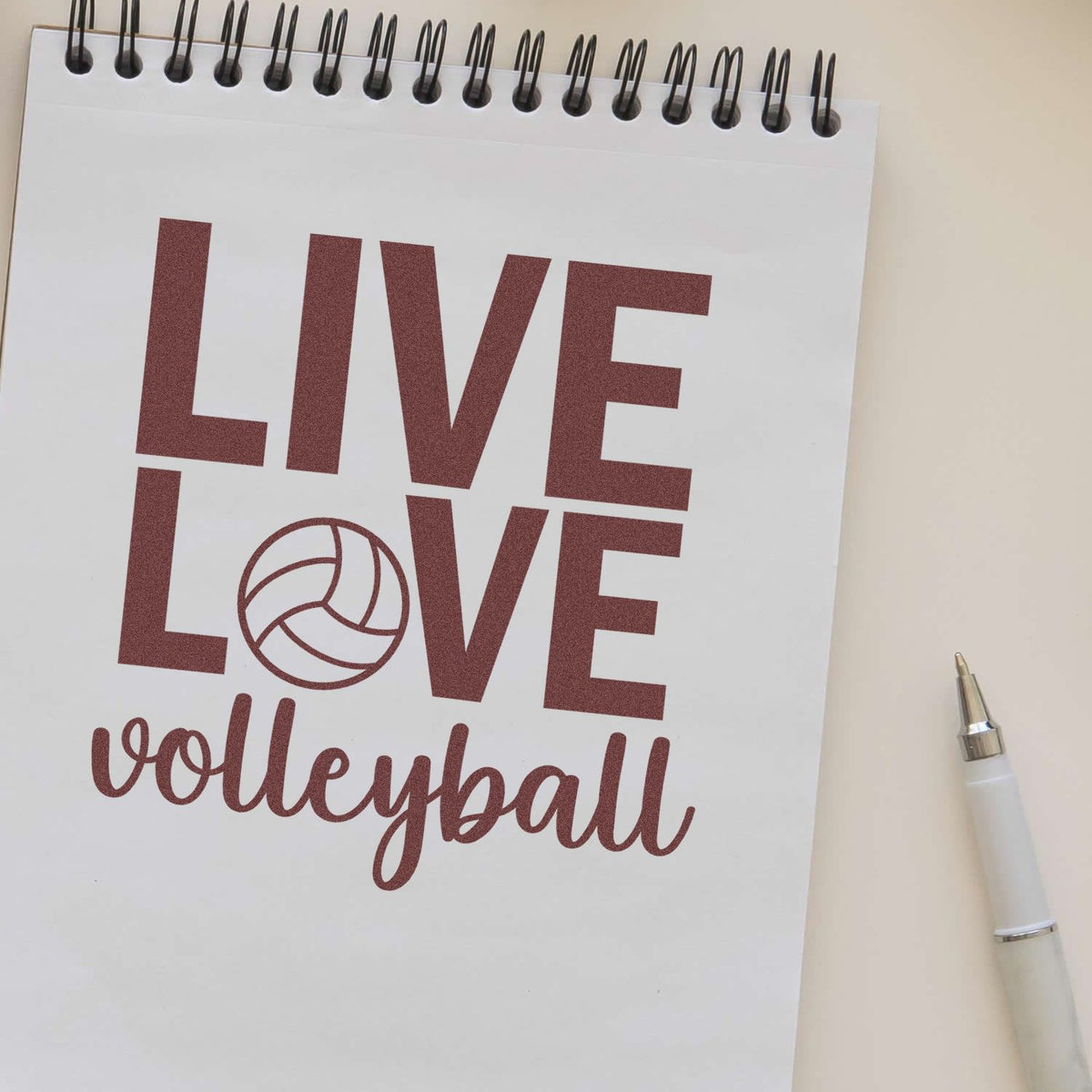 Maroon Live Love volleyball stamp design on white spiral notebook, with volleyball icon and silver pen.