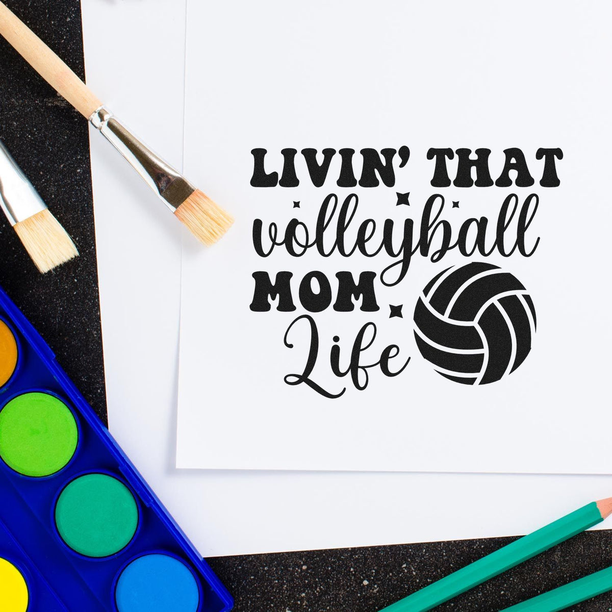 Black volleyball mom life stamp and seal design with ball graphic on white paper, framed by paintbrushes and bright watercolor paints.